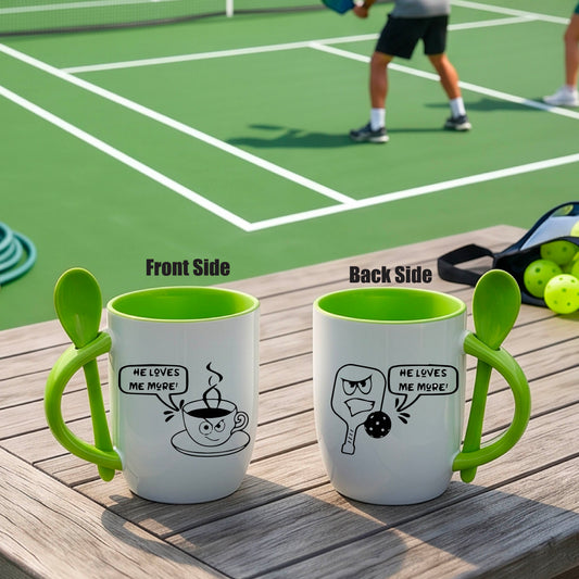 A white and green 12 ounce coffee mug with a built in spoon in the handle with and a fun pickleball paddle and ball and coffee cup graphic and the text “HE LOVES ME MORE”