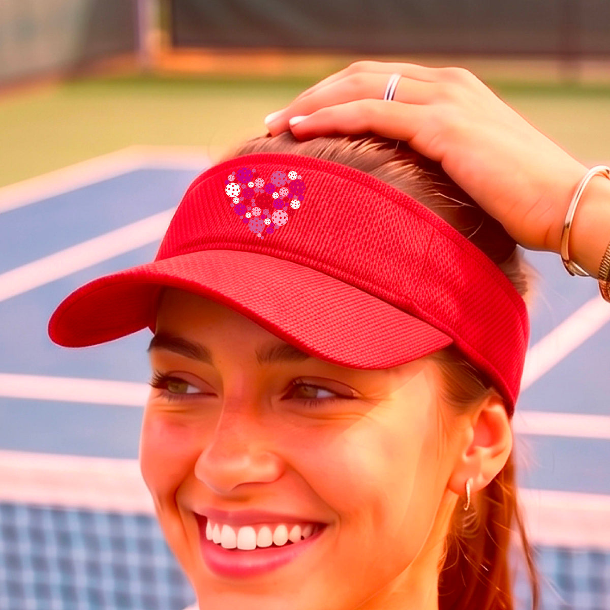 A close-up of a red visor decorated with a heart-shaped pickelball pattern in pink, red, and white graphic. 