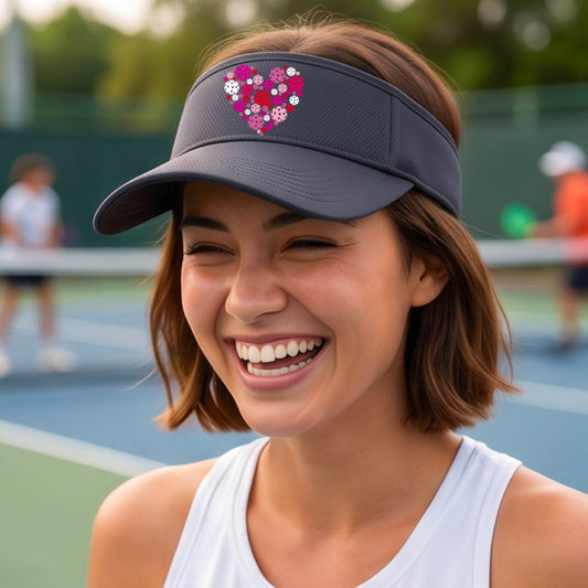 A woman on a pickleball court wearing a dark gray visor decorated with a heart-shaped pickelball pattern in pink, red, and white graphic. 
