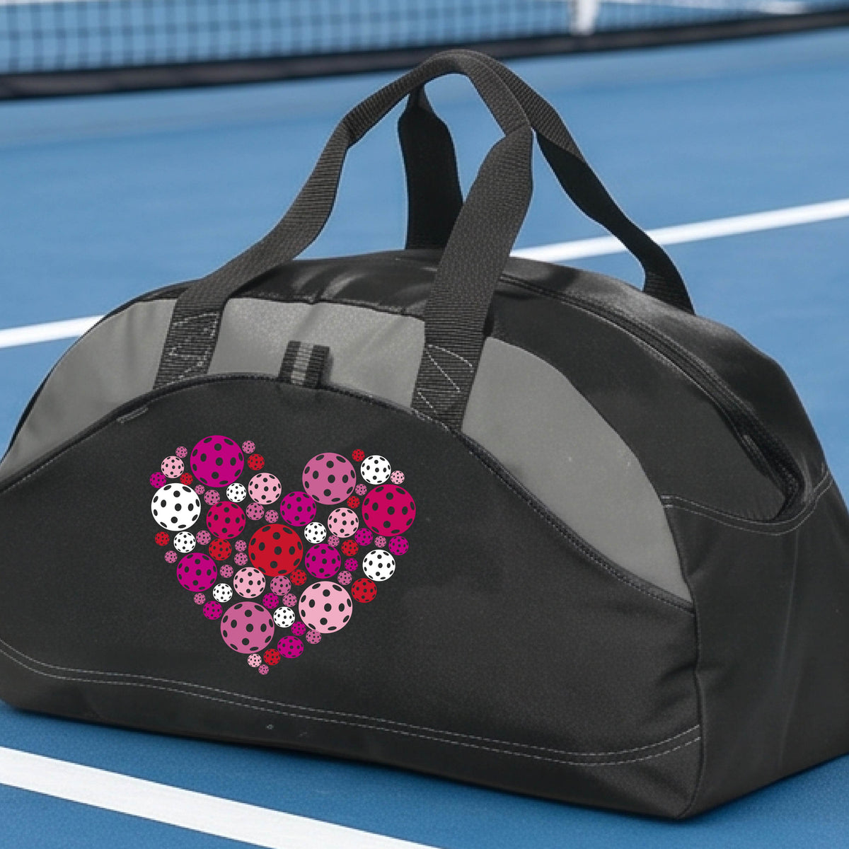 A black duffel bag with gray panels, featuring a heart-shaped pattern of pink, red, and white pickelballs on the front. The bag is shown on a pickleball court in the background.