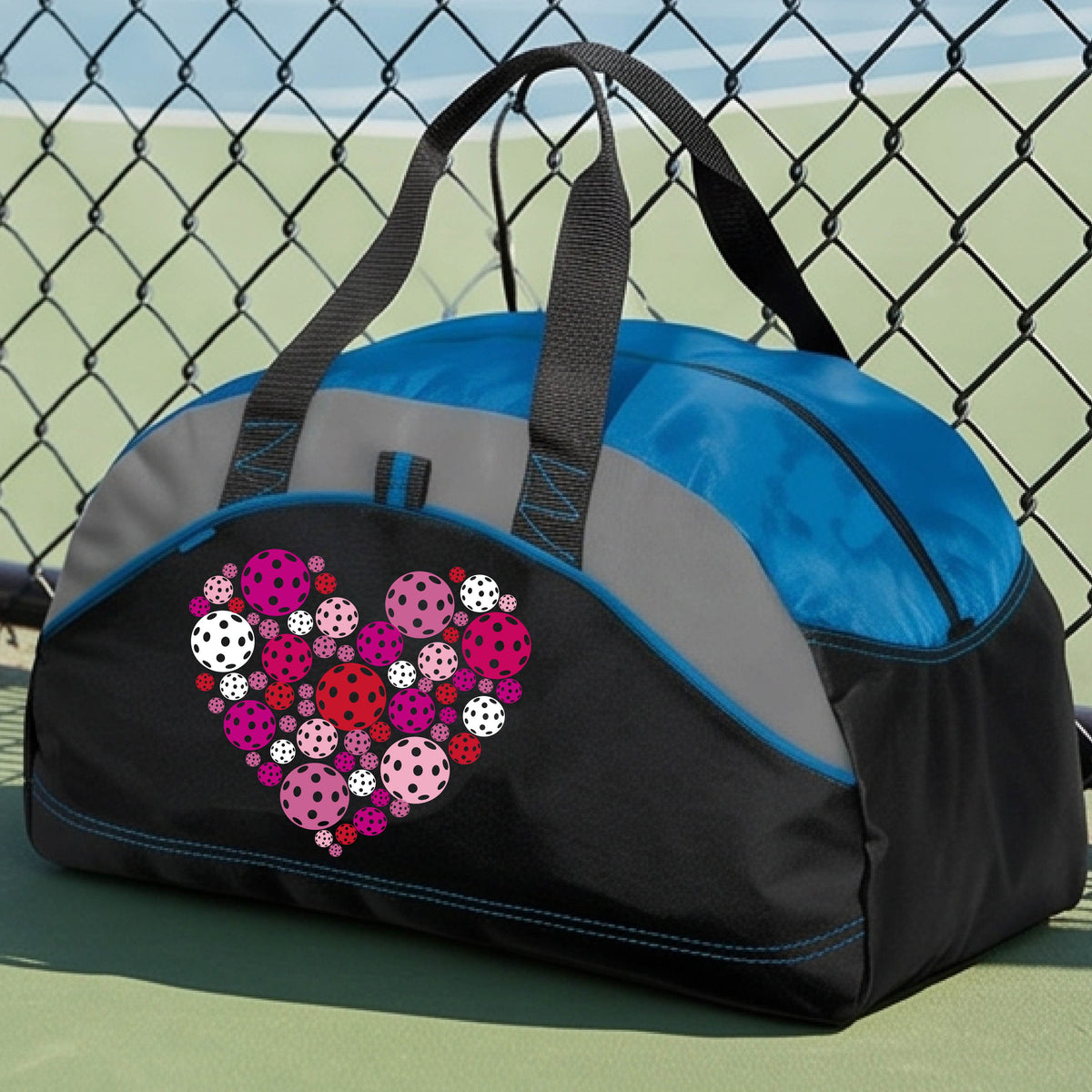 A black and royal blue duffel bag with gray panels, featuring a heart-shaped pattern of pink, red, and white pickelballs on the front. The bag is shown on a pickleball court in the background.