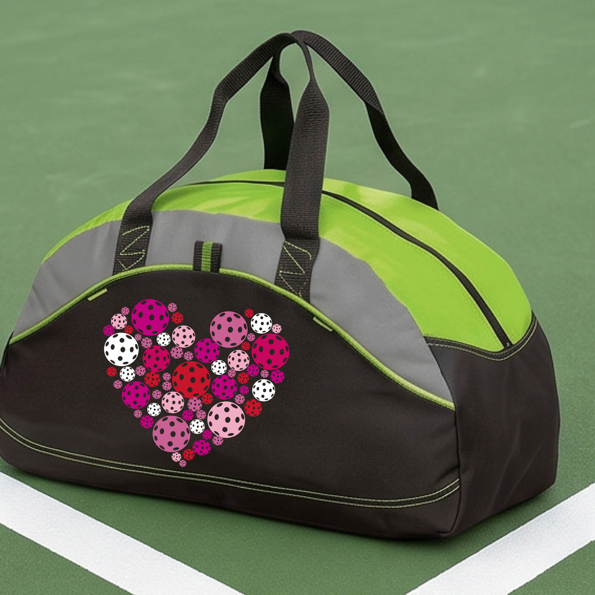 A black and lime green duffel bag with gray panels, featuring a heart-shaped pattern of pink, red, and white pickelballs on the front. The bag is shown on a pickleball court in the background.