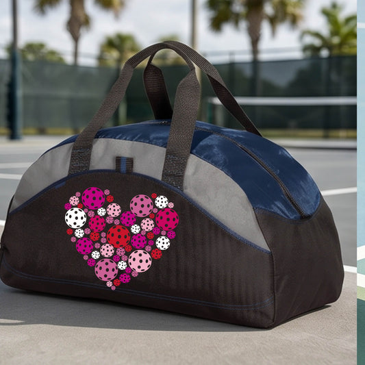A black and navy blue duffel bag with gray panels, featuring a heart-shaped pattern of pink, red, and white pickelballs on the front. The bag is shown on a pickleball court in the background.