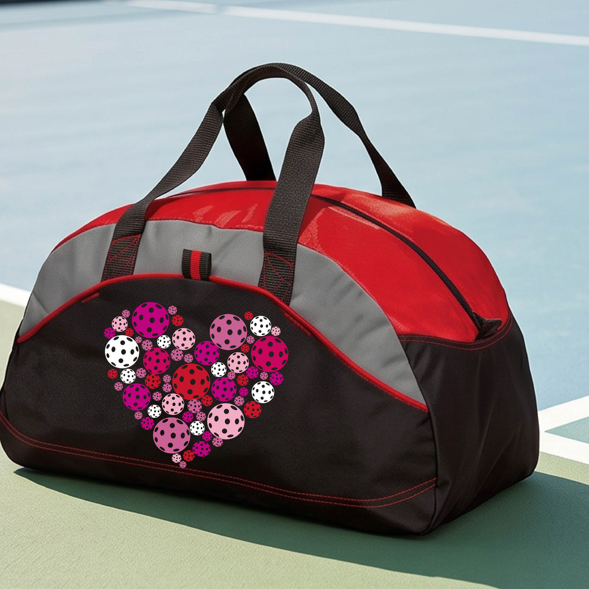 A black and red duffel bag with gray panels, featuring a heart-shaped pattern of pink, red, and white pickelballs on the front. The bag is shown on a pickleball court in the background.