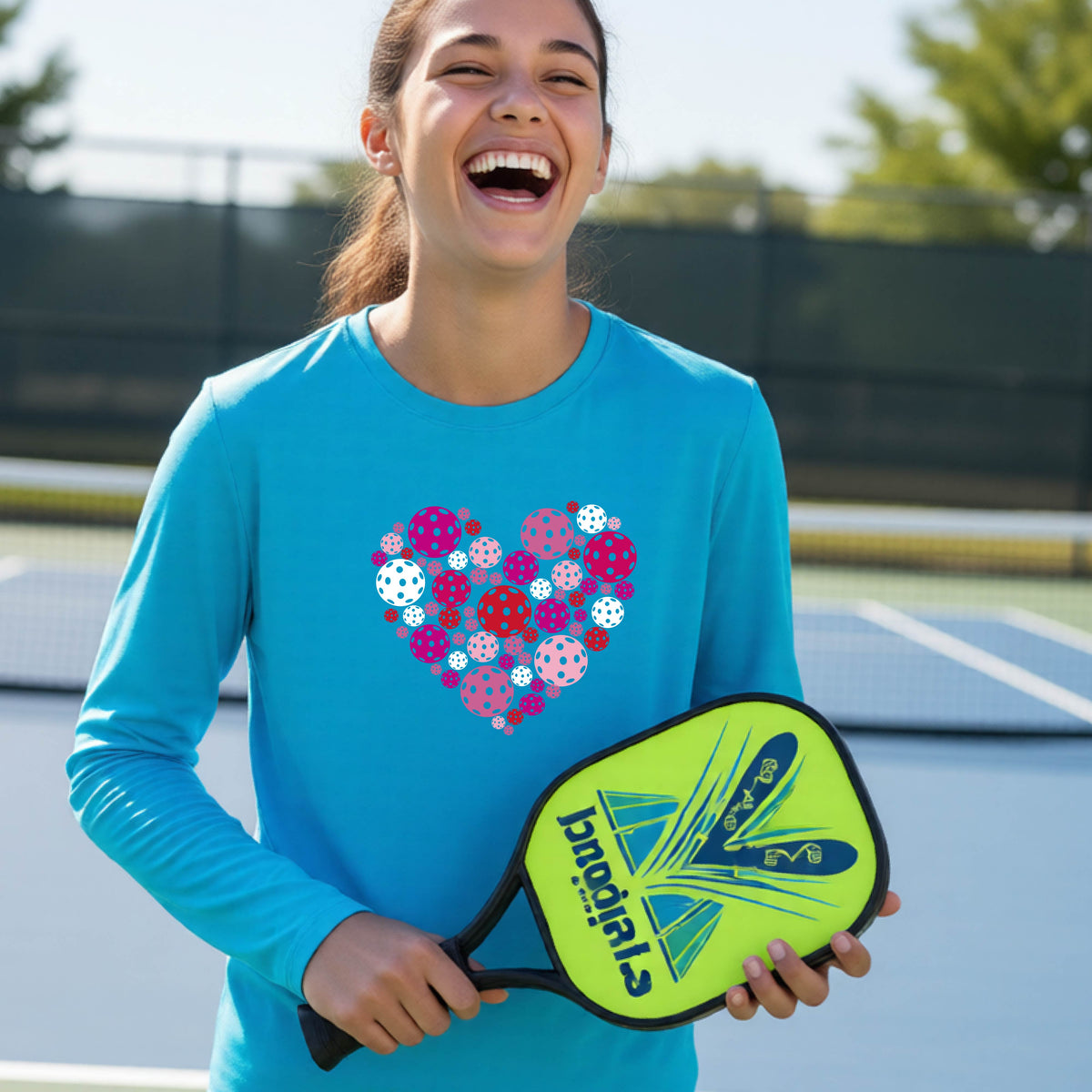 A teeneager laughing on a pickleball court wearing a bright blue long-sleeve shirt with a colorful heart-shaped graphic made of pickleballs. 