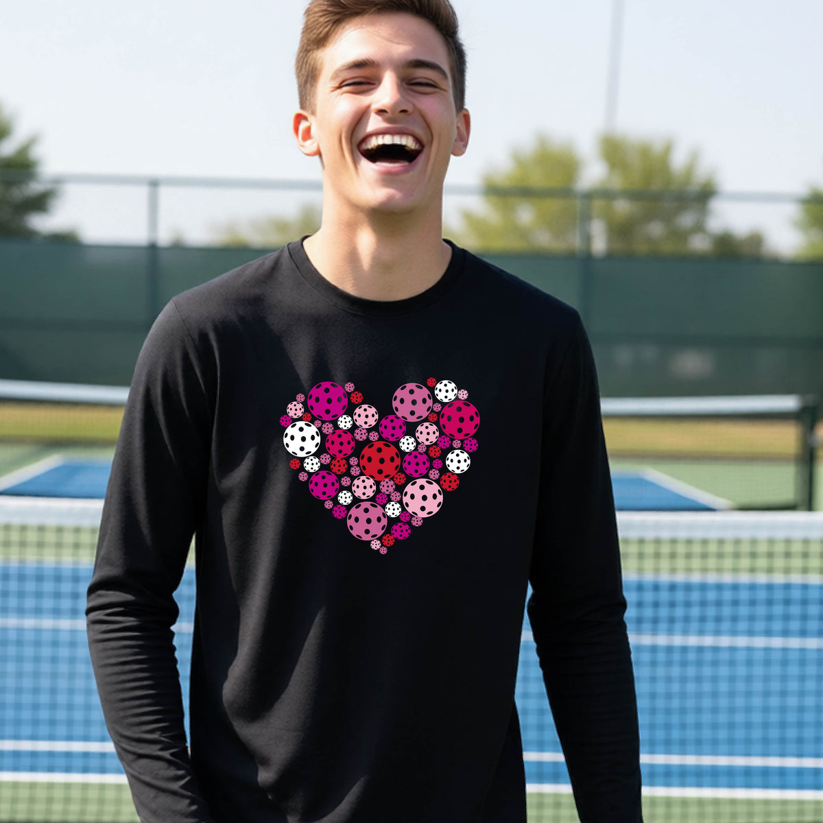 A teeneager laughing on a pickleball court wearing a black long-sleeve shirt with a colorful heart-shaped graphic made of pickleballs. 