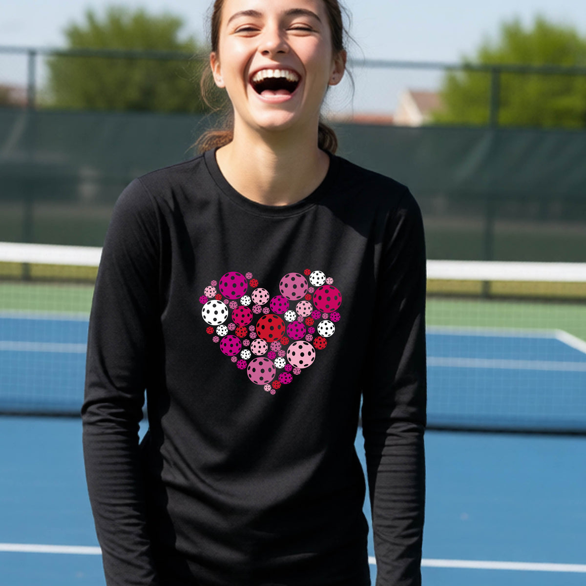 A teeneager laughing on a pickleball court wearing a black long-sleeve shirt with a colorful heart-shaped graphic made of pickleballs. 