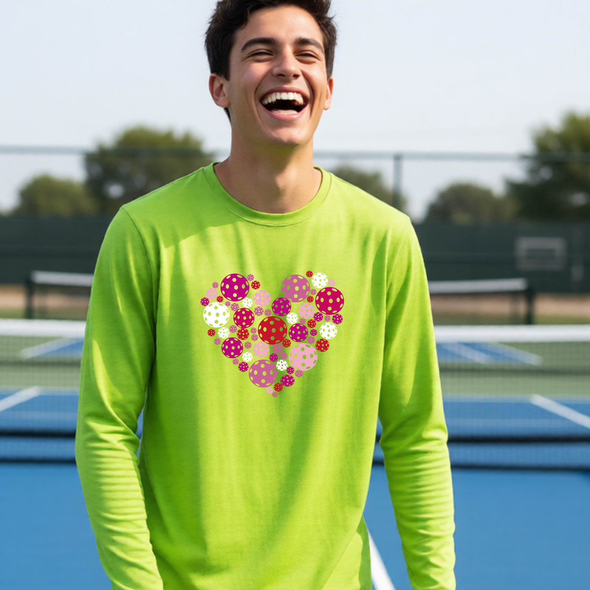 A teeneager laughing on a pickleball court wearing a lime green long-sleeve shirt with a colorful heart-shaped graphic made of pickleballs. 