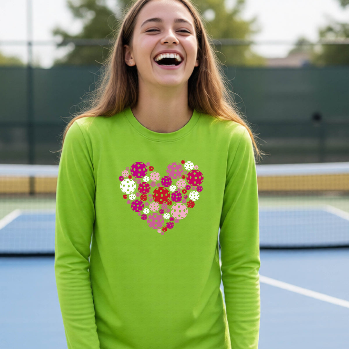 A teeneager laughing on a pickleball court wearing a lime green long-sleeve shirt with a colorful heart-shaped graphic made of pickleballs. 