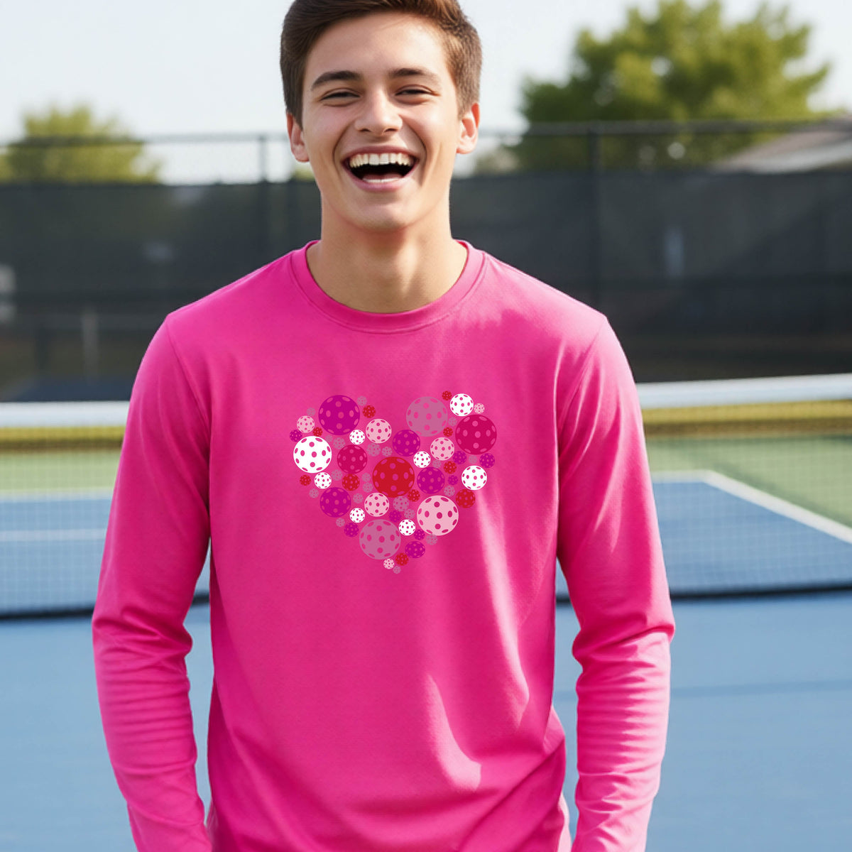 A teeneager laughing on a pickleball court wearing a bright pink long-sleeve shirt with a colorful heart-shaped graphic made of pickleballs. 