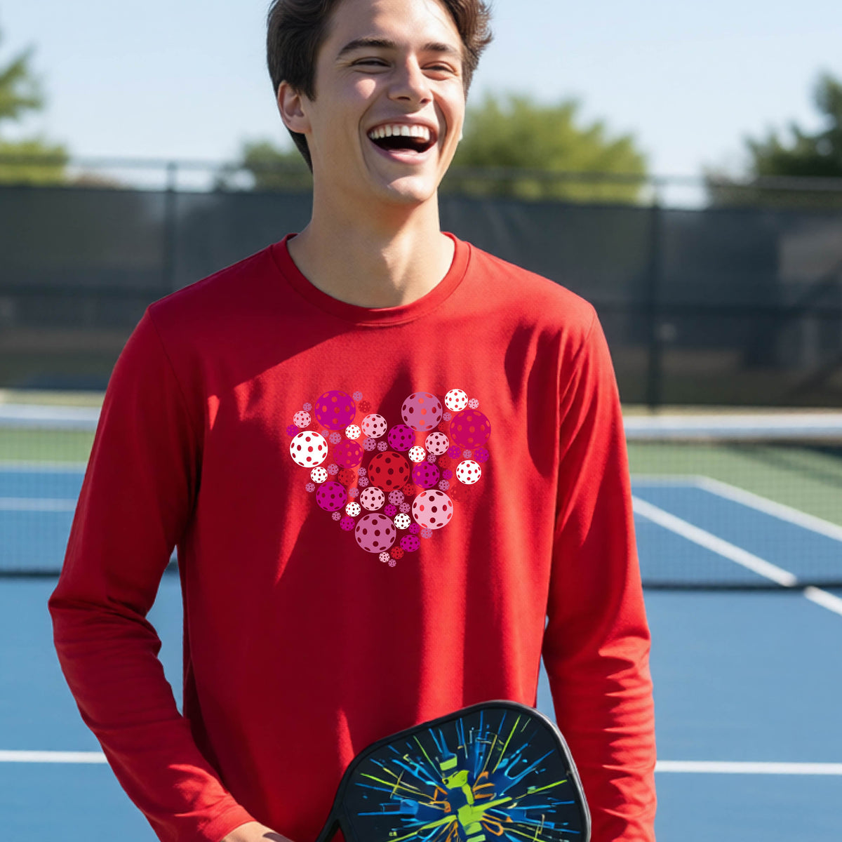 A teeneager laughing on a pickleball court wearing a red long-sleeve shirt with a colorful heart-shaped graphic made of pickleballs. 