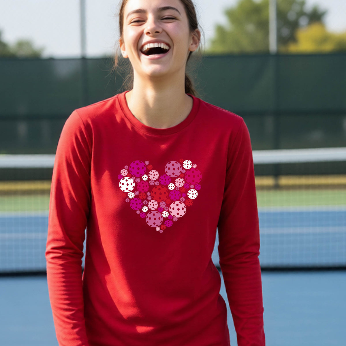 A teeneager laughing on a pickleball court wearing a red long-sleeve shirt with a colorful heart-shaped graphic made of pickleballs. 