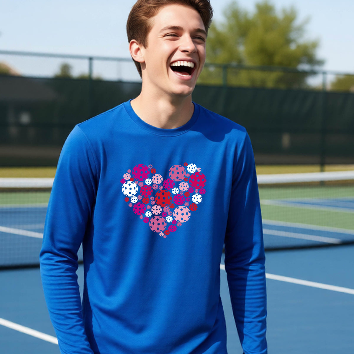 A teeneager laughing on a pickleball court wearing a royal blue long-sleeve shirt with a colorful heart-shaped graphic made of pickleballs. 