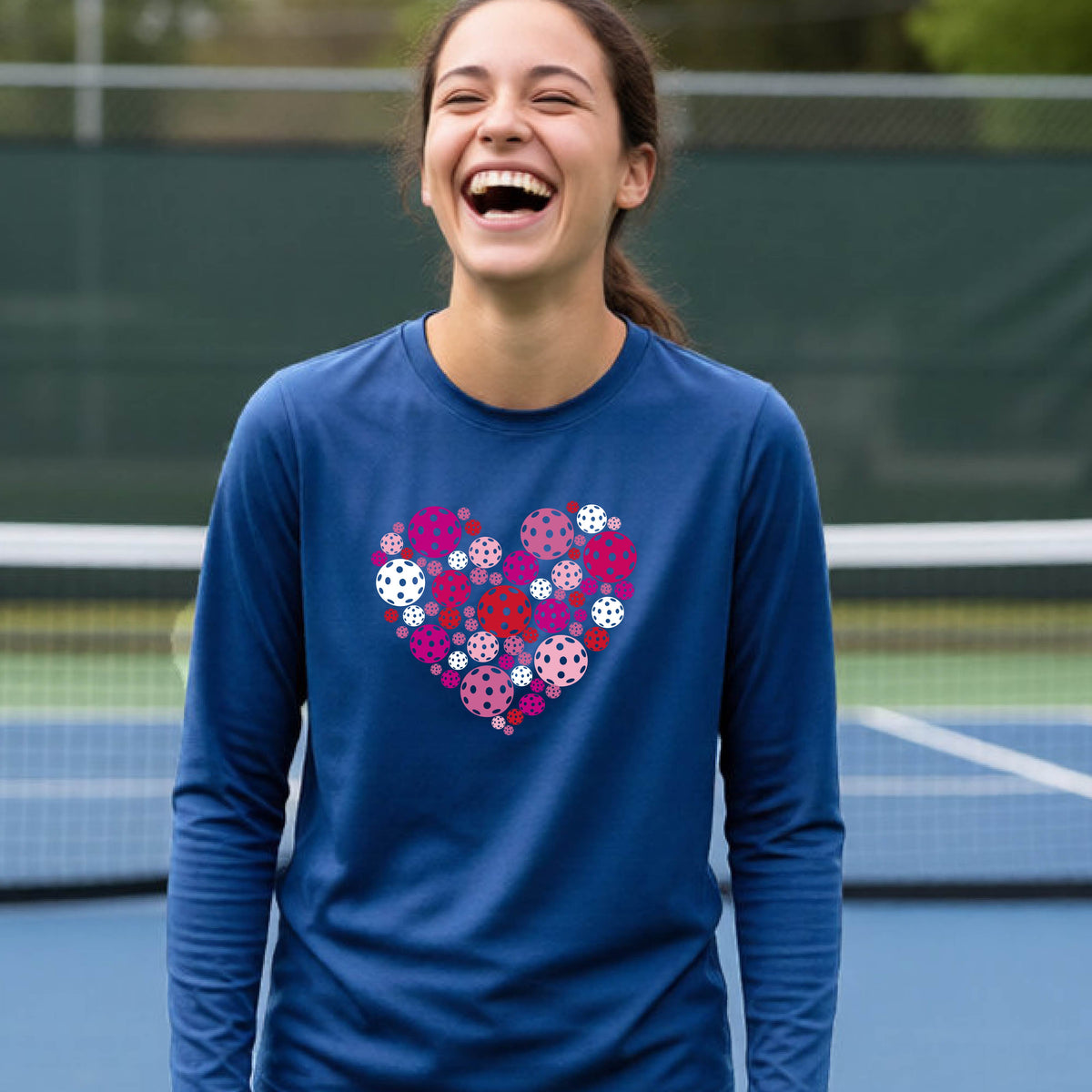 A teeneager laughing on a pickleball court wearing a royal blue long-sleeve shirt with a colorful heart-shaped graphic made of pickleballs. 