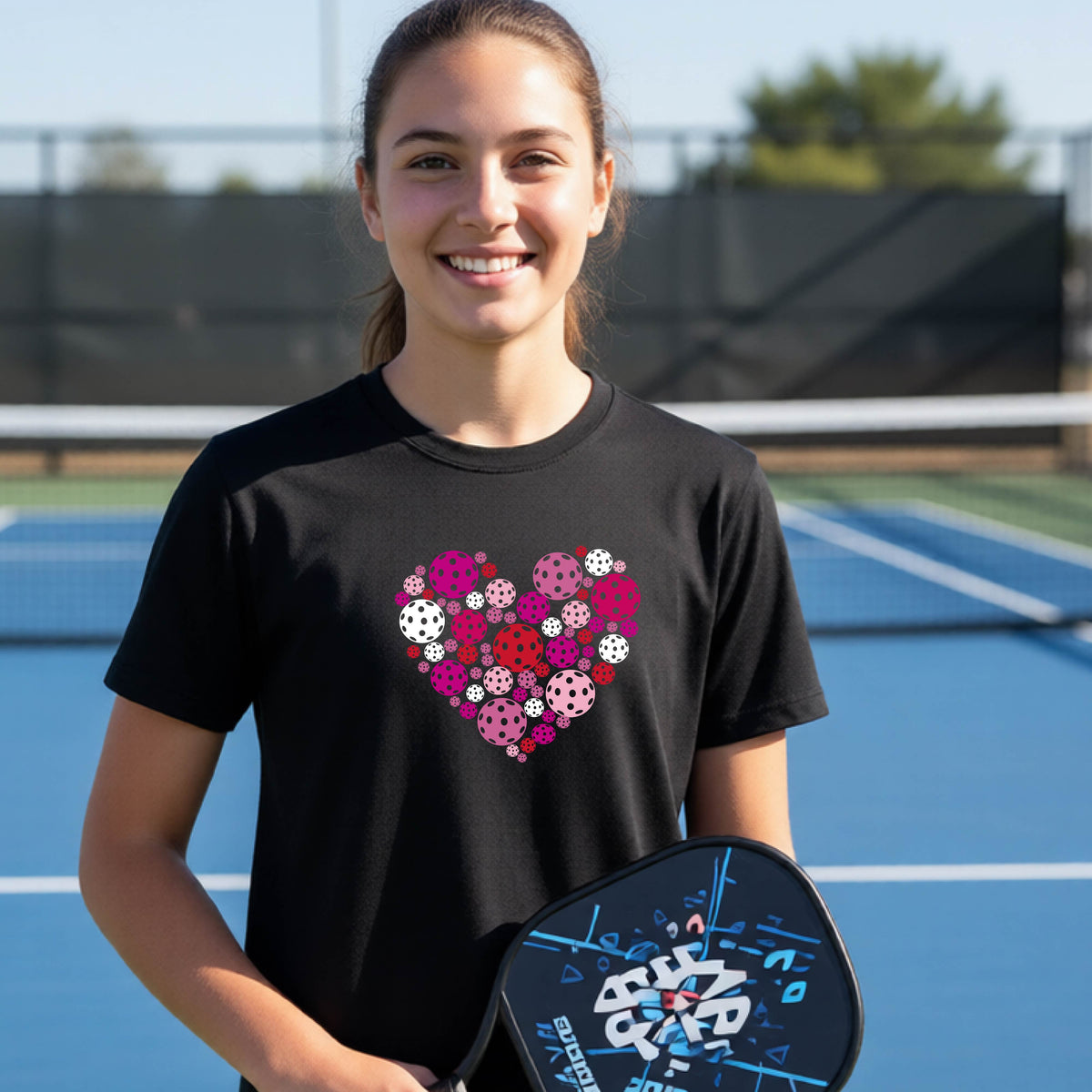 A young woman laughing on an outdoor pickleball court wearing a black t-shirt with a heart-shaped graphic made of pink, red, and white pickleballs. 