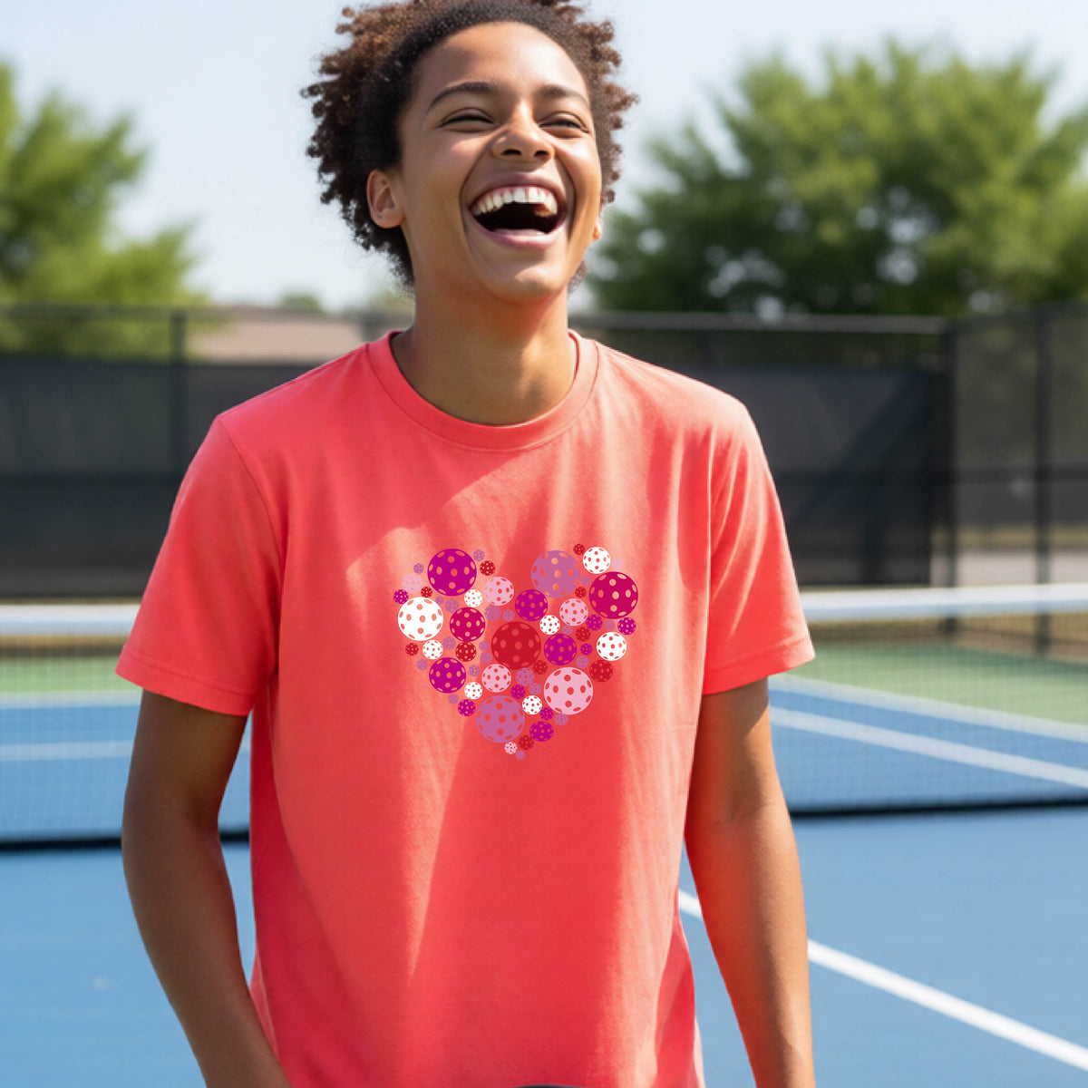 A young person laughing on an outdoor pickleball court wearing a coral pink t-shirt with a heart-shaped graphic made of pink, red, and white pickleballs. 