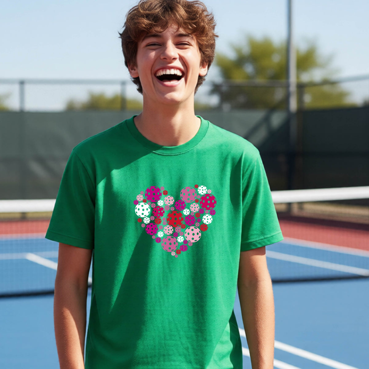 A young person laughing on an outdoor pickleball court wearing a green t-shirt with a heart-shaped graphic made of pink, red, and white pickleballs. 