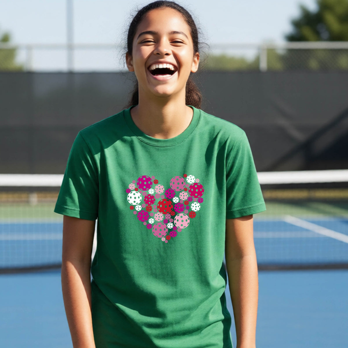 A young person laughing on an outdoor pickleball court wearing a green t-shirt with a heart-shaped graphic made of pink, red, and white pickleballs. 