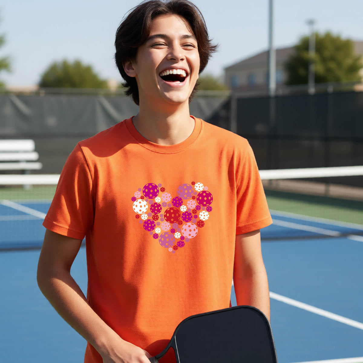 A young person laughing on an outdoor pickleball court wearing an orange t-shirt with a heart-shaped graphic made of pink, red, and white pickleballs. 