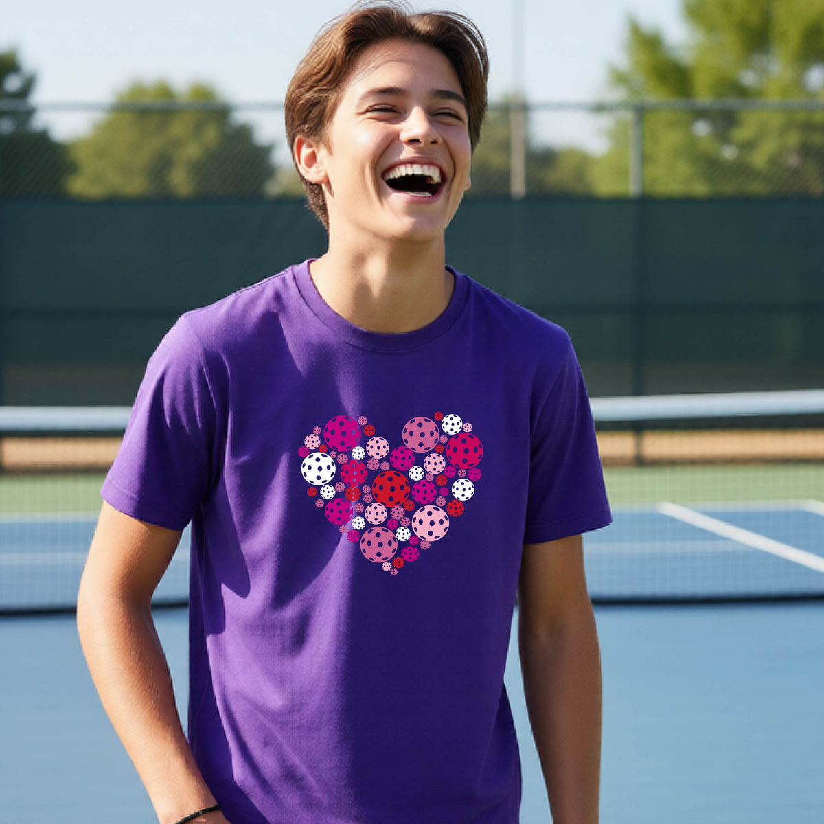 A young person laughing on an outdoor pickleball court wearing a purple t-shirt with a heart-shaped graphic made of pink, red, and white pickleballs. 