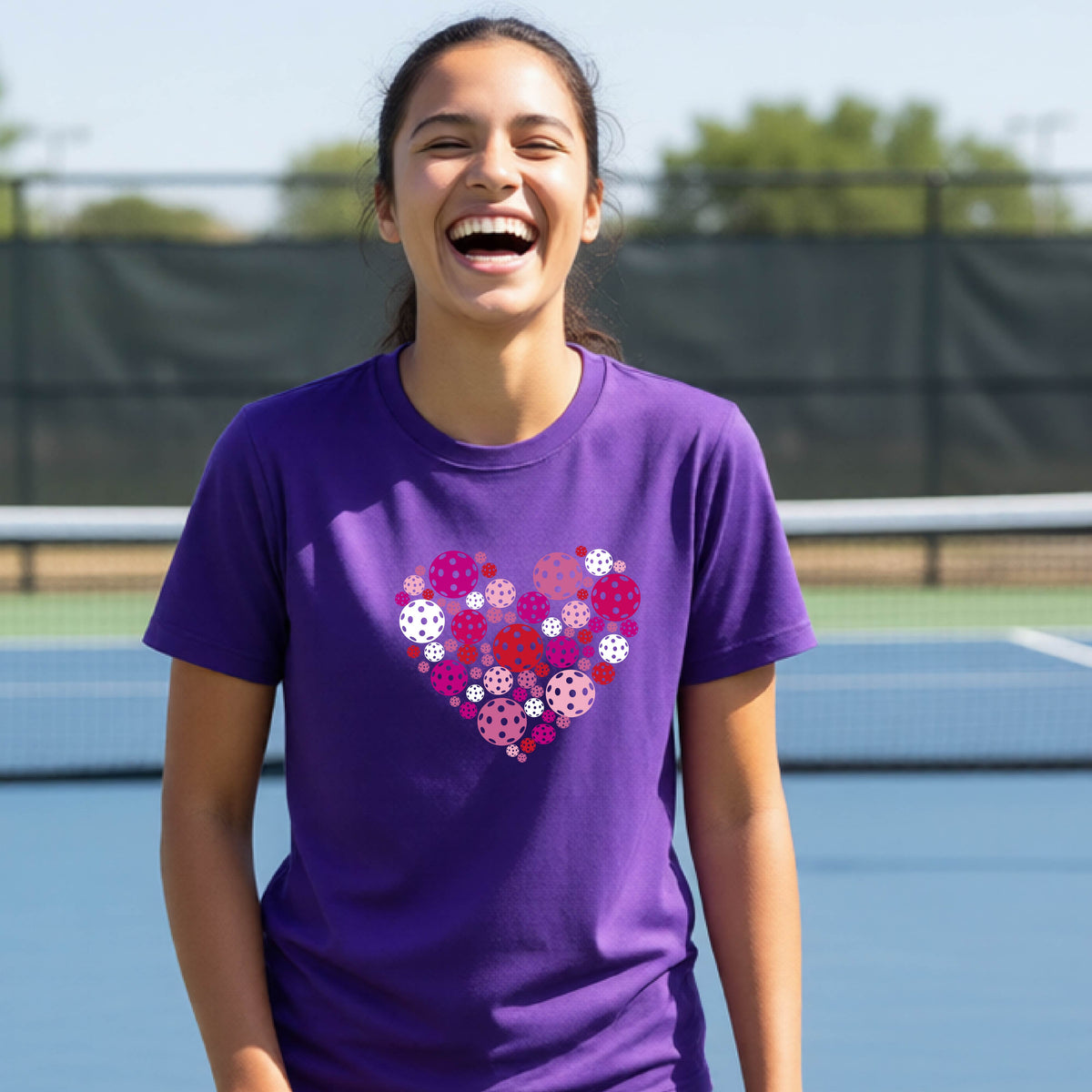 A woman laughing on an outdoor pickleball court wearing a purple t-shirt with a heart-shaped graphic made of pink, red, and white pickleballs. 
