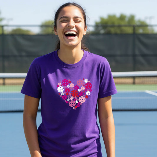 A woman laughing on an outdoor pickleball court wearing a purple t-shirt with a heart-shaped graphic made of pink, red, and white pickleballs. 