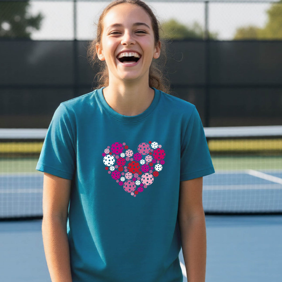A young woman laughing on an outdoor pickleball court wearing a teal t-shirt with a heart-shaped graphic made of pink, red, and white pickleballs. 