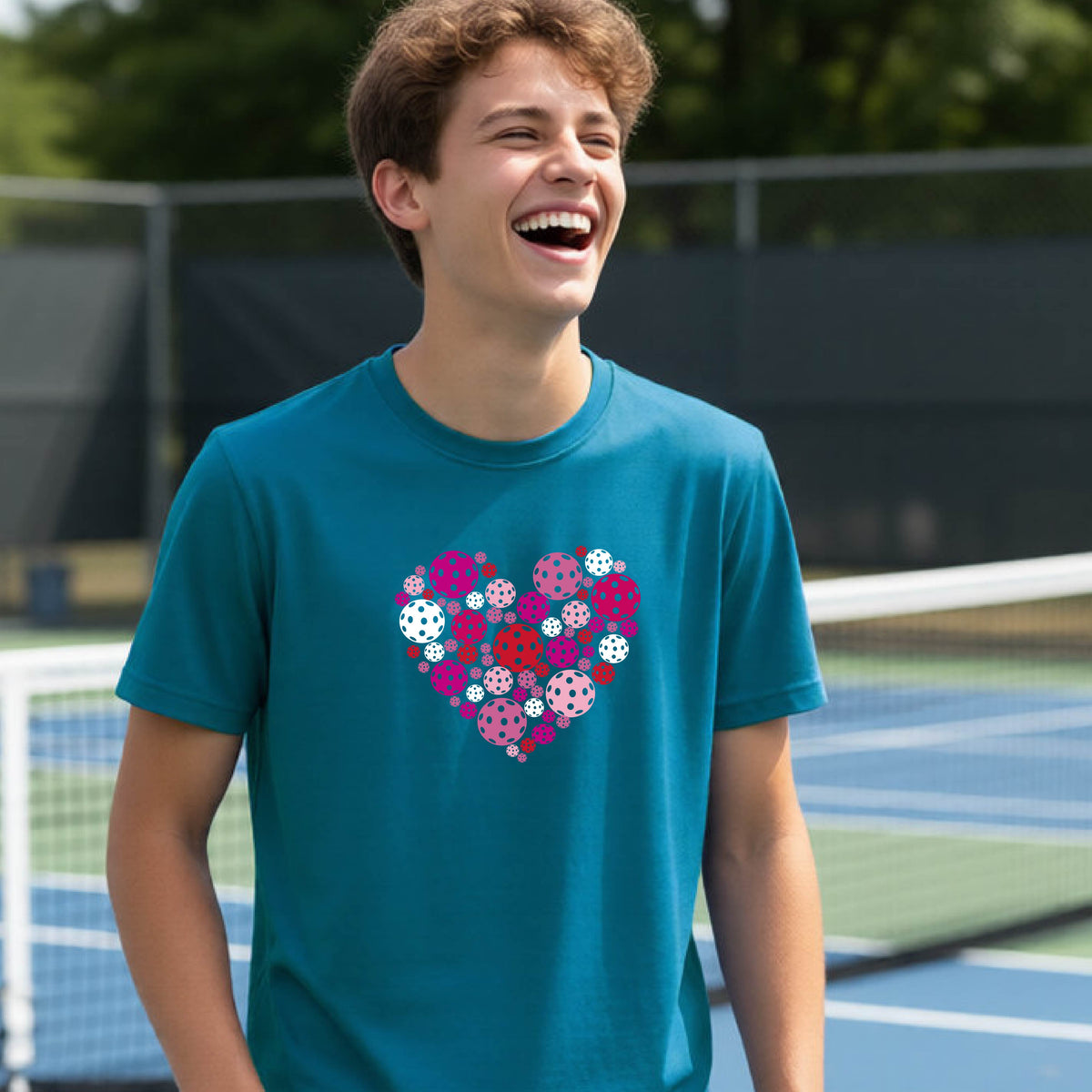 A young person laughing on an outdoor pickleball court wearing a teal t-shirt with a heart-shaped graphic made of pink, red, and white pickleballs. 