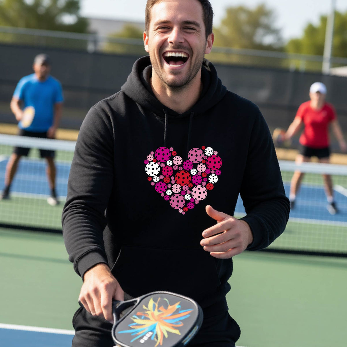 A smiling man on a pickleball court wearing a black hoodie with a heart-shaped graphic made of colorful pickleballls.