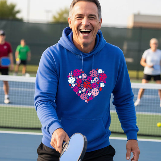 A smiling man on a pickleball court wearing a bright blue hoodie with a heart-shaped graphic made of colorful pickleballls.