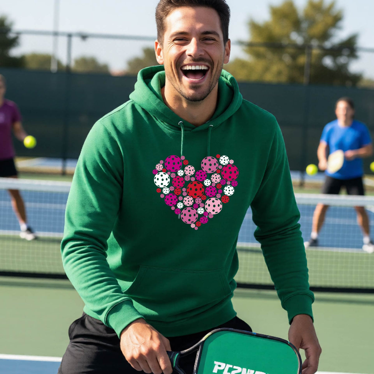 A smiling man on a pickleball court wearing a green hoodie with a heart-shaped graphic made of colorful pickleballls.