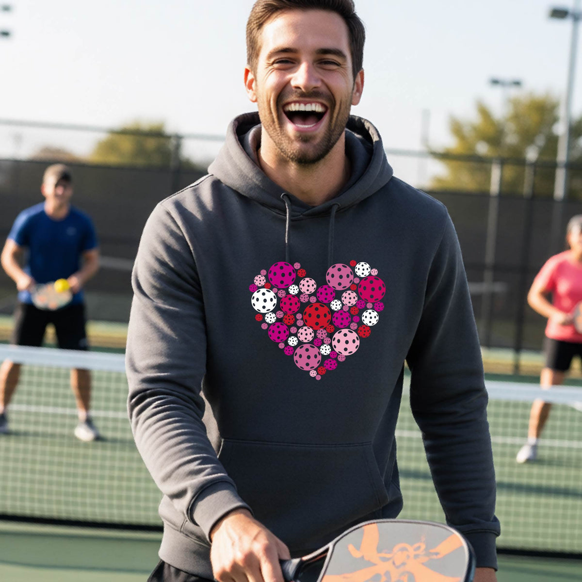 A smiling man on a pickleball court wearing a gray hoodie with a heart-shaped graphic made of colorful pickleballls.