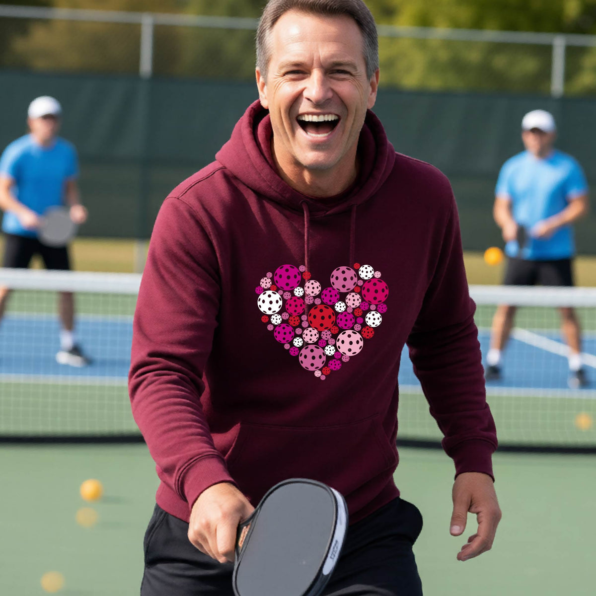 A smiling man on a pickleball court wearing a maroon hoodie with a heart-shaped graphic made of colorful pickleballls.