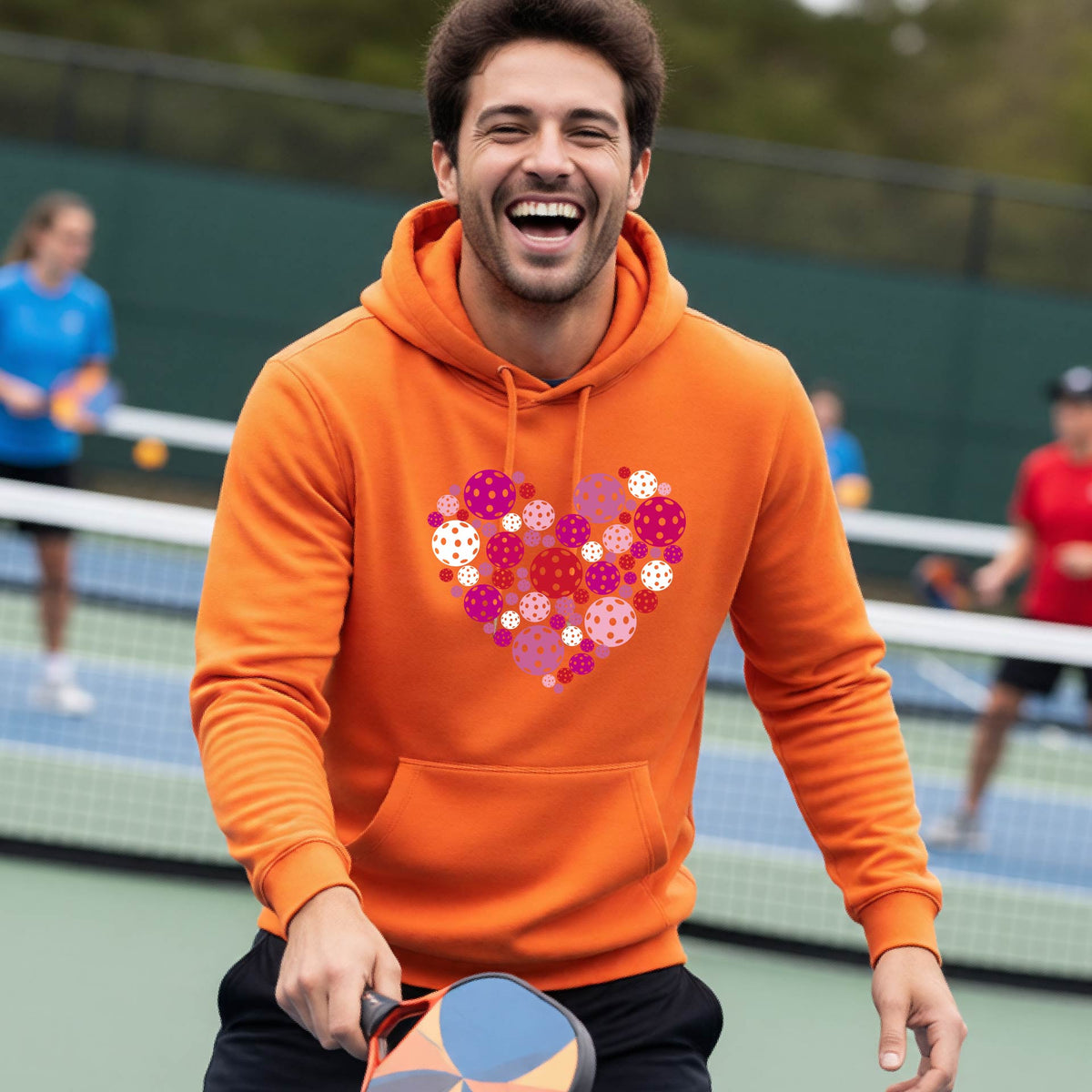 A smiling man on a pickleball court wearing a bright orange hoodie with a heart-shaped graphic made of colorful pickleballls.