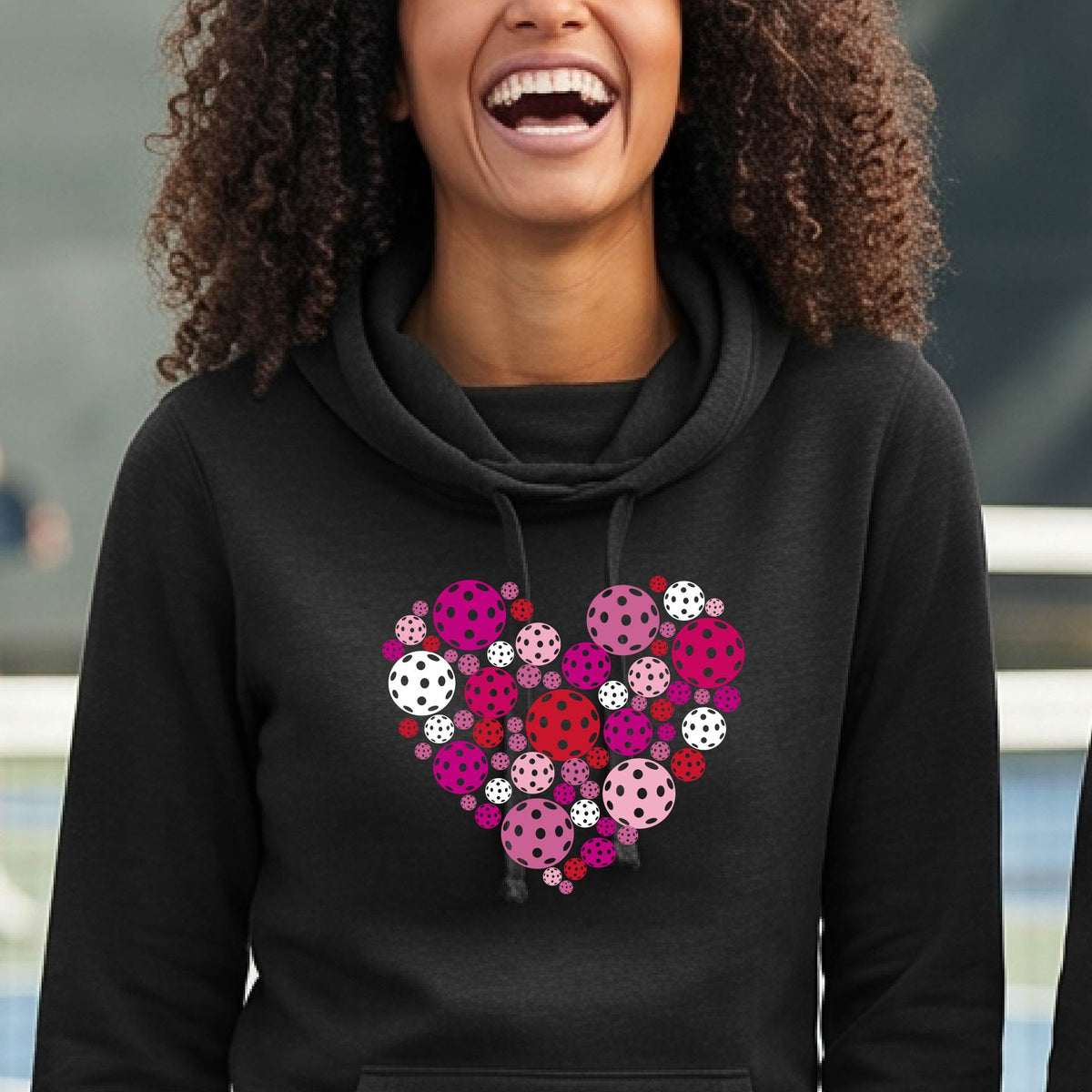 A woman wearing a black cowl-neck hoodie with a colorful heart-shaped pickleball pattern on the chest, set against a blurred outdoor background.