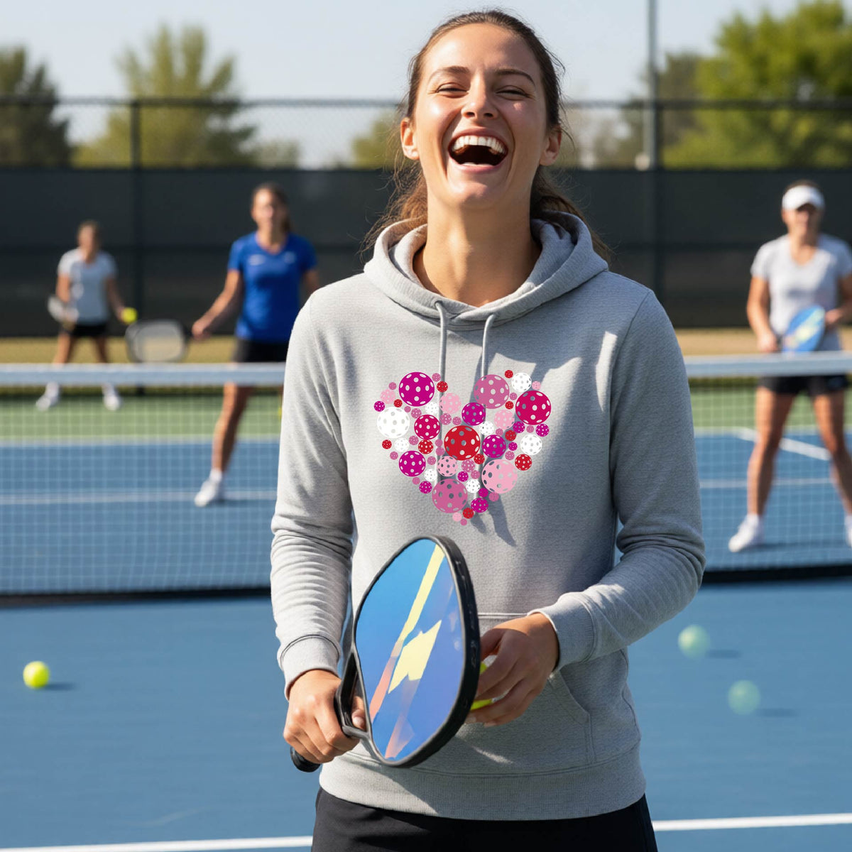 A woman wearing a gray cowl-neck hoodie with a colorful heart-shaped pickleball pattern on the chest, set against a blurred outdoor background.