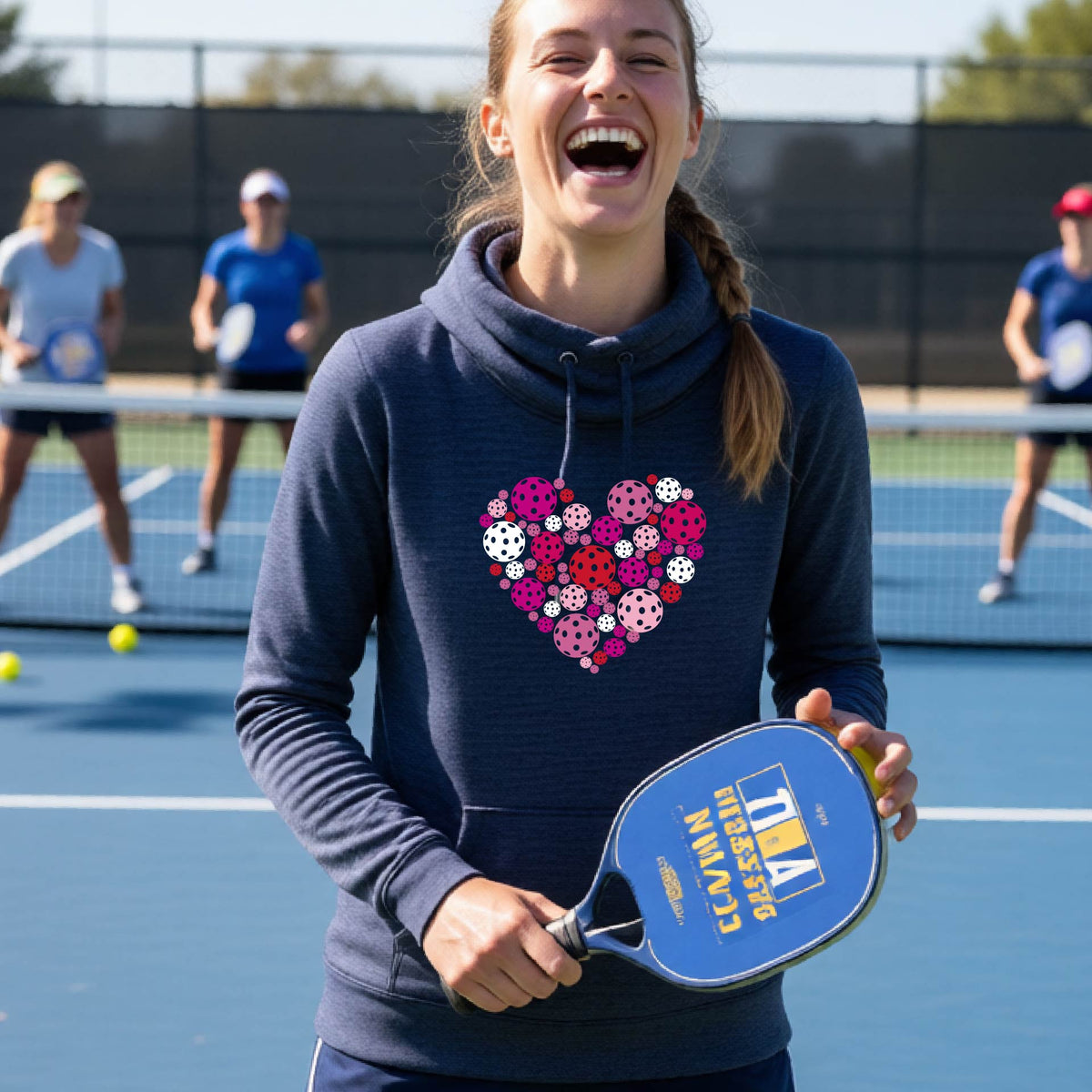 A woman wearing a navy blue cowl-neck hoodie with a colorful heart-shaped pickleball pattern on the chest, set against a blurred outdoor background.