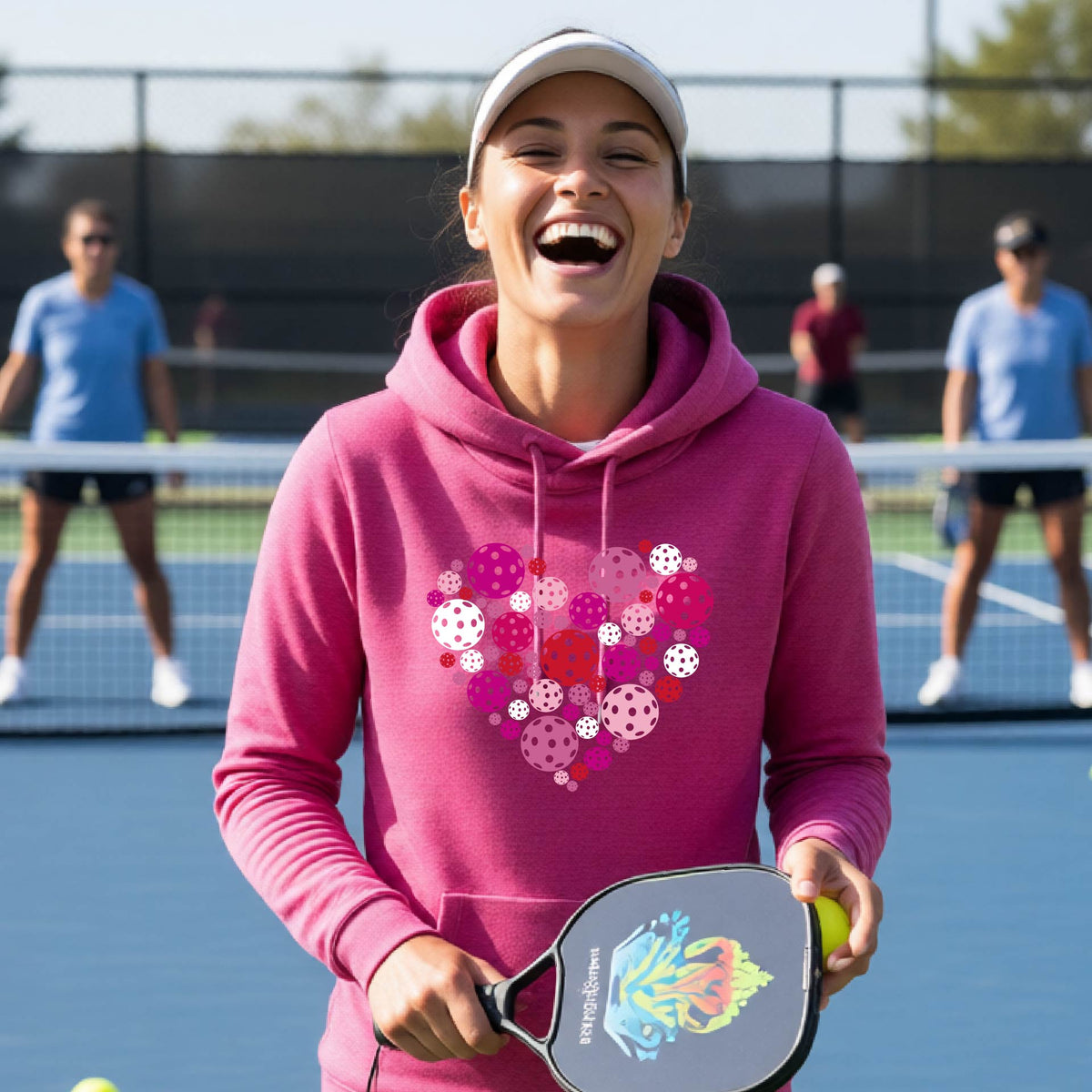 A woman wearing a pink cowl-neck hoodie with a colorful heart-shaped pickleball pattern on the chest, set against a blurred outdoor background.