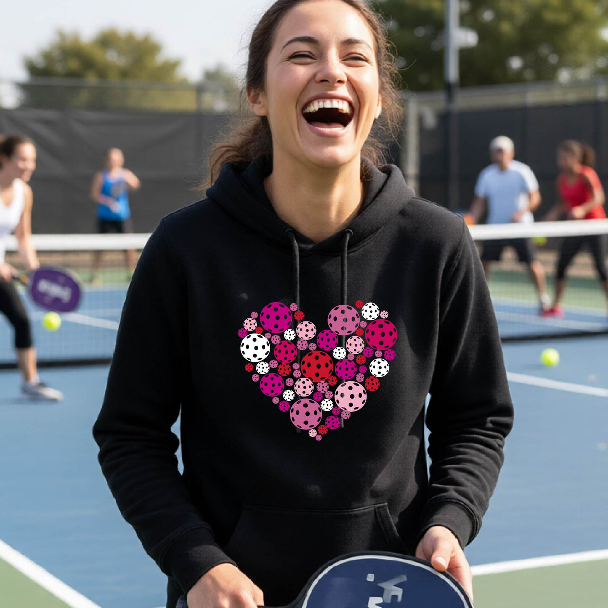 A woman laughing on a pickleball court wearing a black hoodie with a heart-shaped graphic made of pink, red, and white pickleballs. 