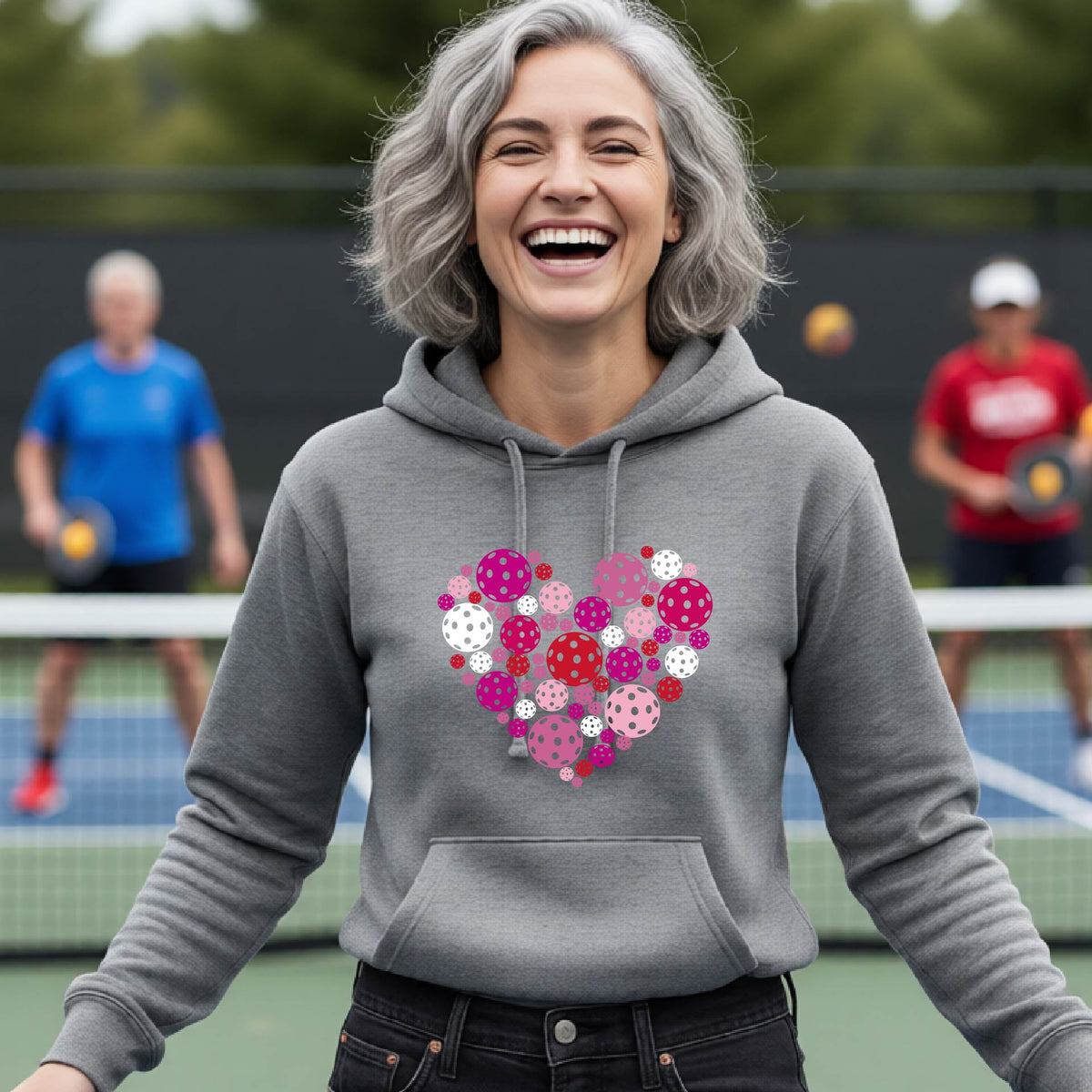 A woman laughing on a pickleball court wearing a gray hoodie with a heart-shaped graphic made of pink, red, and white pickleballs. 