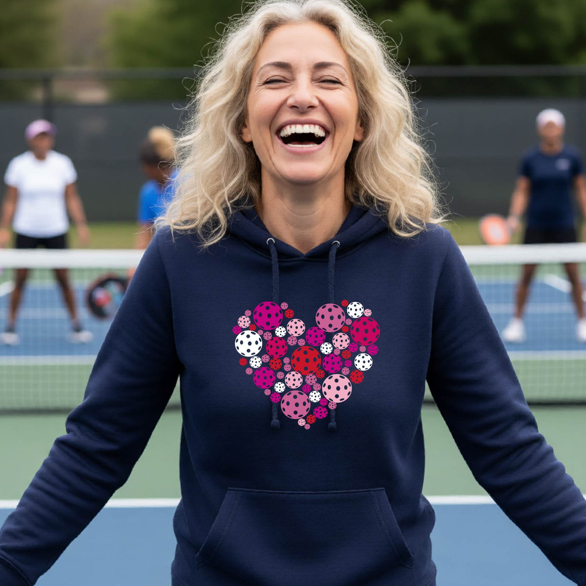 A woman laughing on a pickleball court wearing a navy blue hoodie with a heart-shaped graphic made of pink, red, and white pickleballs. 