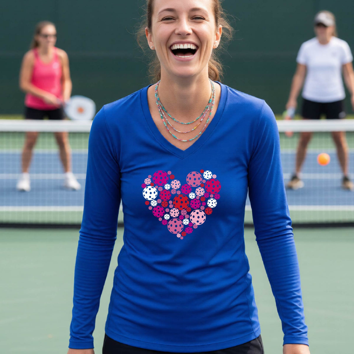 A woman on a pickleball court wearing a royal blue long-sleeve V-neck shirt with a heart-shaped graphic made of pink, red, purple, and white pickleballs on the chest.