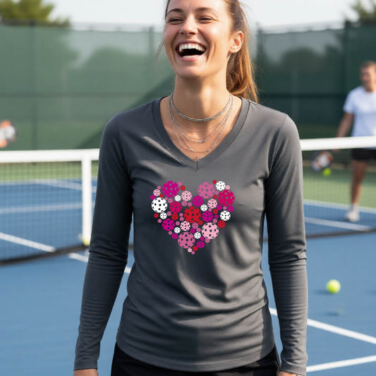 A woman on a pickleball court wearing a dark gray long-sleeve V-neck shirt with a heart-shaped graphic made of pink, red, purple, and white pickleballs on the chest.