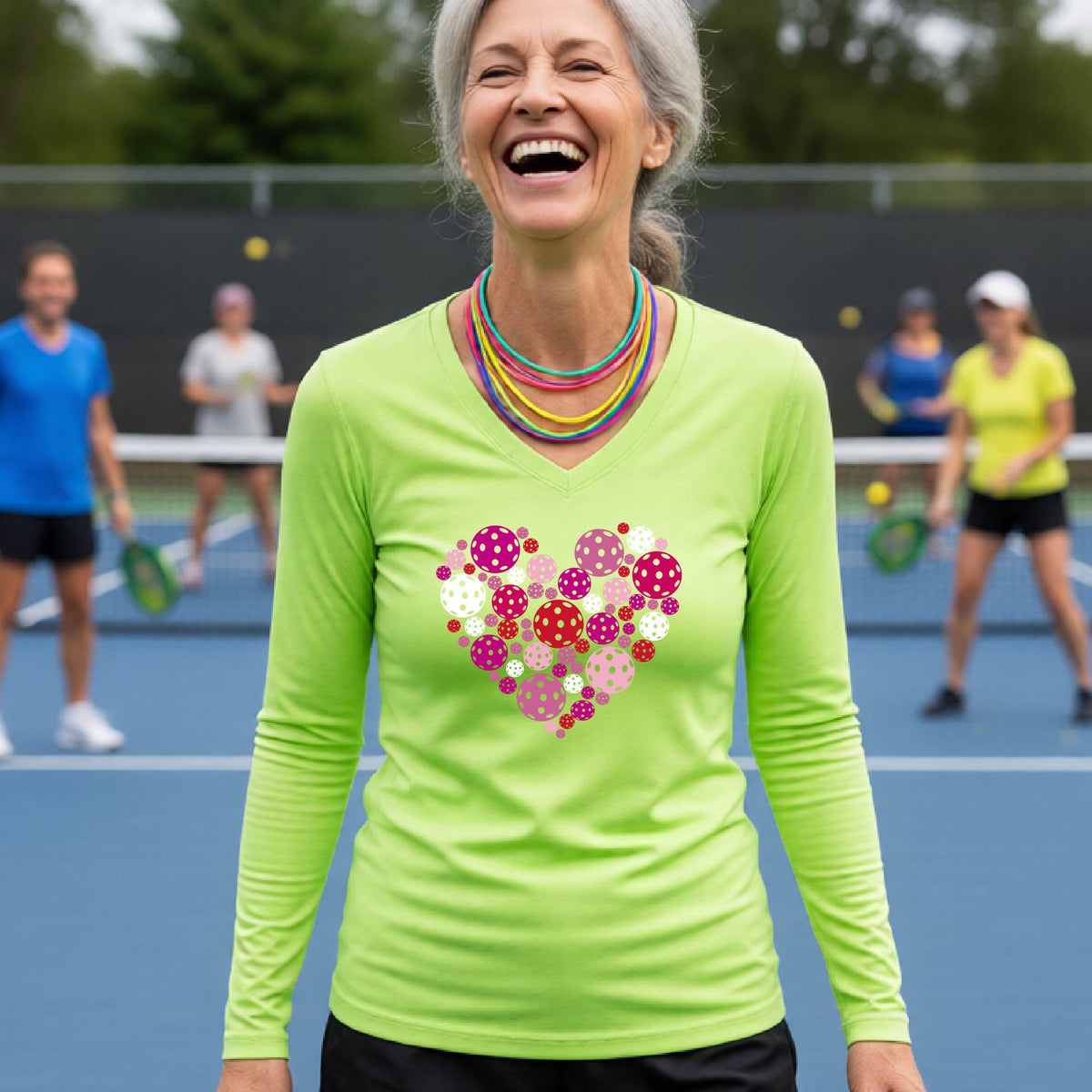 A woman on a pickleball court wearing a lime green long-sleeve V-neck shirt with a heart-shaped graphic made of pink, red, purple, and white pickleballs on the chest.