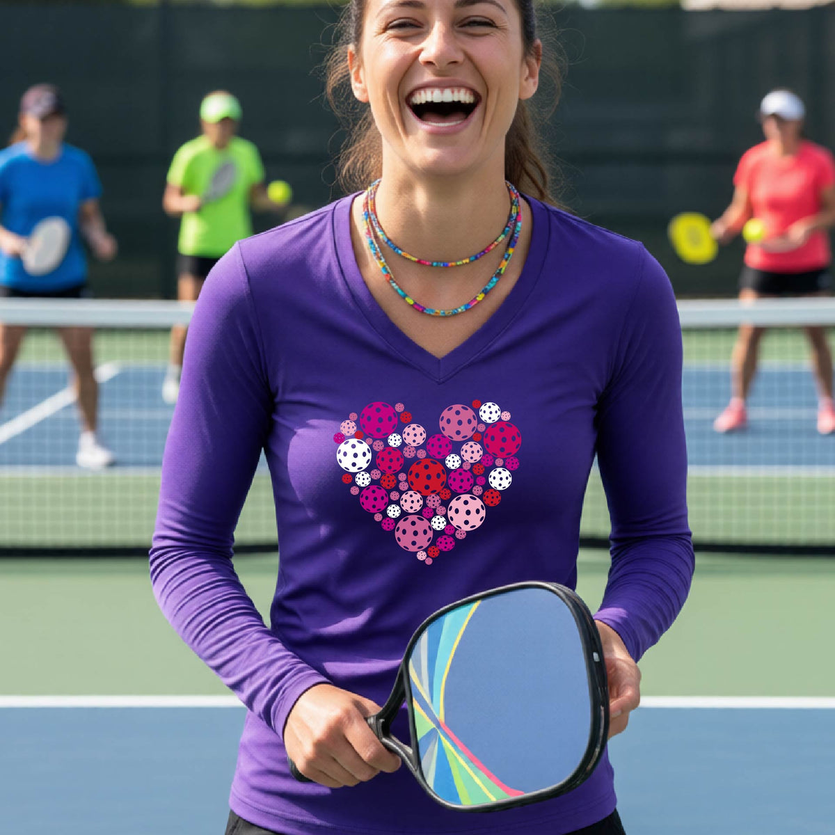 A woman on a pickleball court wearing a purple long-sleeve V-neck shirt with a heart-shaped graphic made of pink, red, purple, and white pickleballs on the chest.