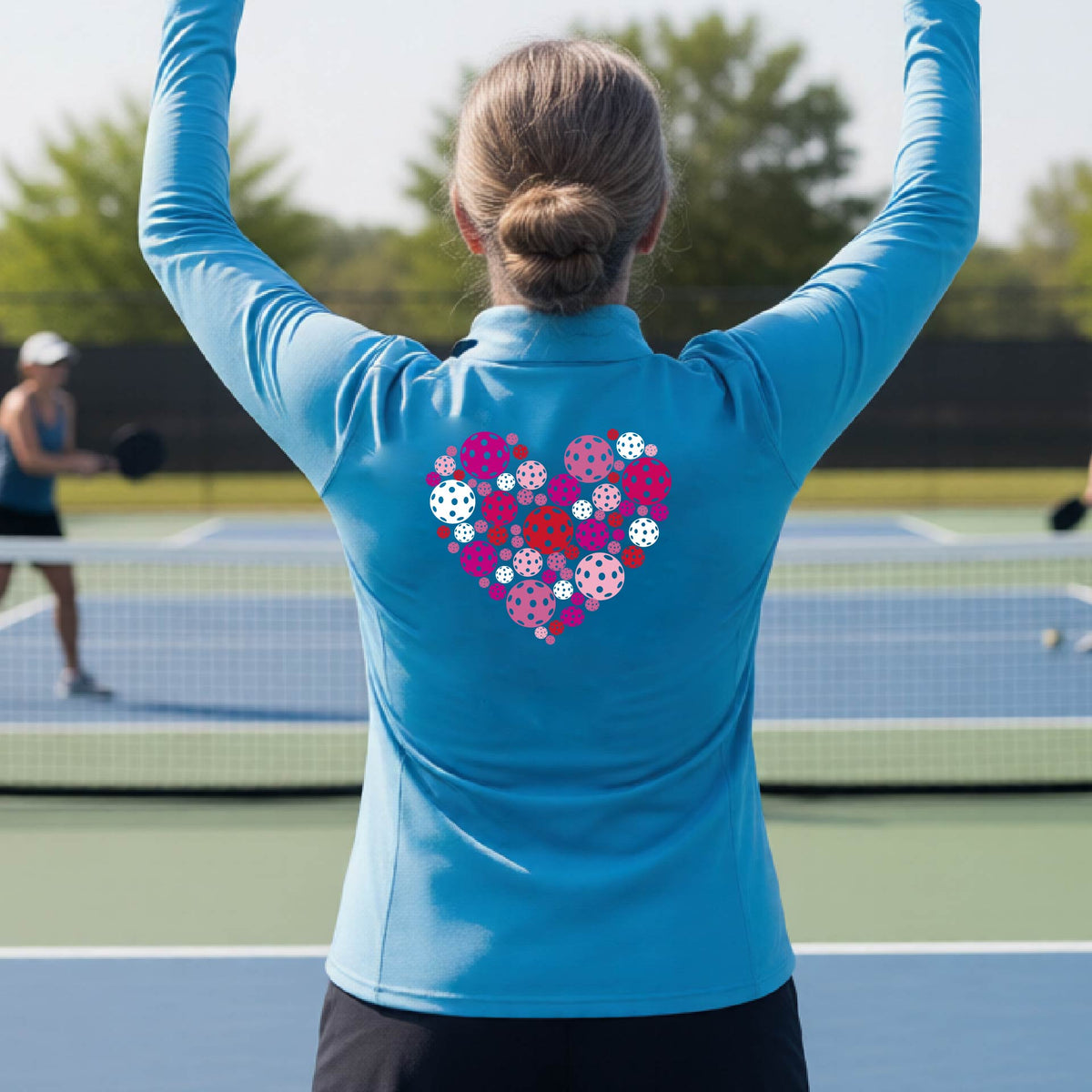 A woman laughing on an outdoor pickleball court wearing a bright blue quarter-zip shirt with a heart-shaped graphic made of pink, red, and white pickleballs on the back. 