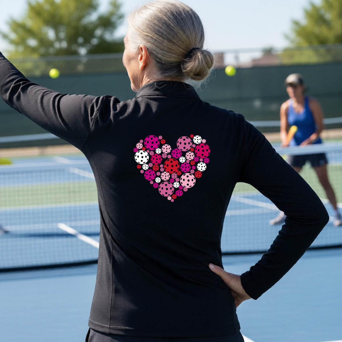 A woman laughing on a pickleball court wearing a black quarter-zip shirt with a colorful heart-shaped pickelball pattern on the chest.