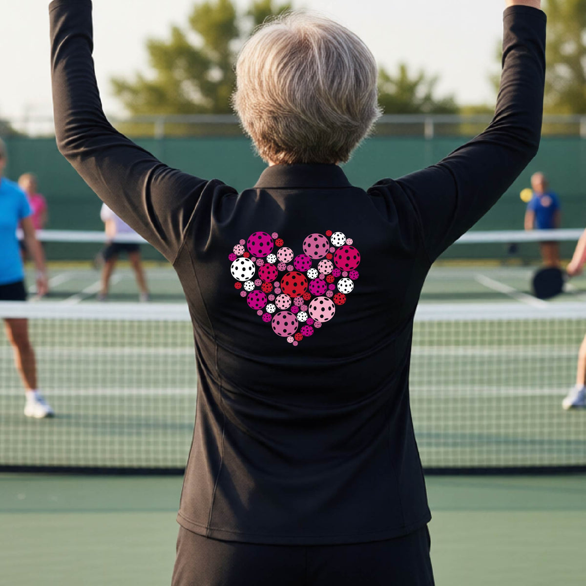 A woman laughing on an outdoor pickleball court wearing a black quarter-zip shirt with a heart-shaped graphic made of pink, red, and white pickleballs on the back. 