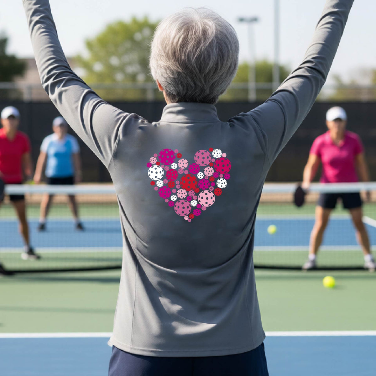 A woman laughing on an outdoor pickleball court wearing a gray quarter-zip shirt with a heart-shaped graphic made of pink, red, and white pickleballs on the back. 