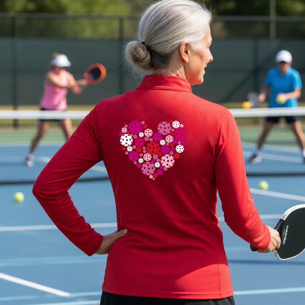 A woman laughing on an outdoor pickleball court wearing a red quarter-zip shirt with a heart-shaped graphic made of pink, red, and white pickleballs on the back. 
